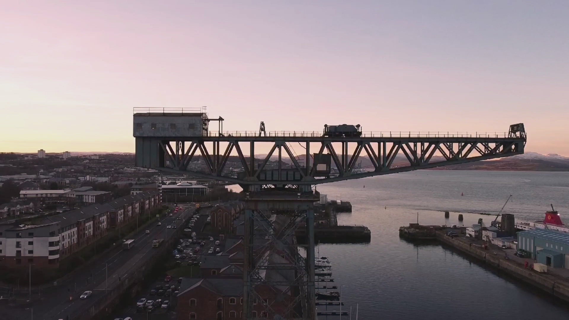 Industrial crane on a bridge above a river at sunset, with buildings and vehicles visible along the waterfront.